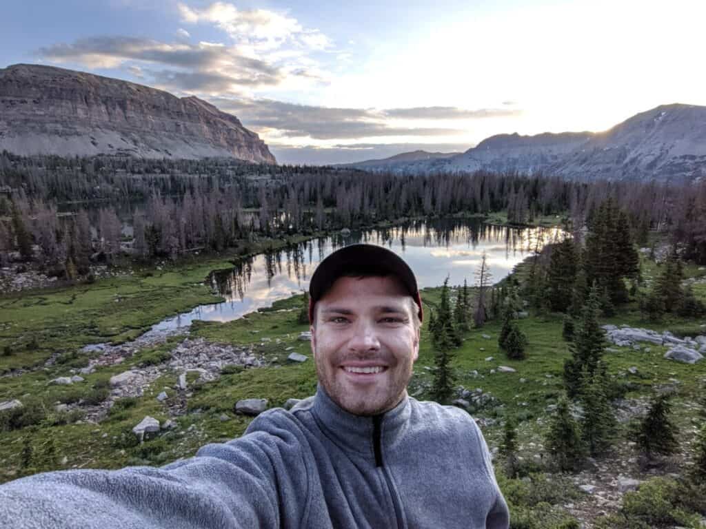 me-wearing-a-fleece-jacket-and-a-baseball-cap-facing-camera-with-mountains-and-mountain-lake-in-background